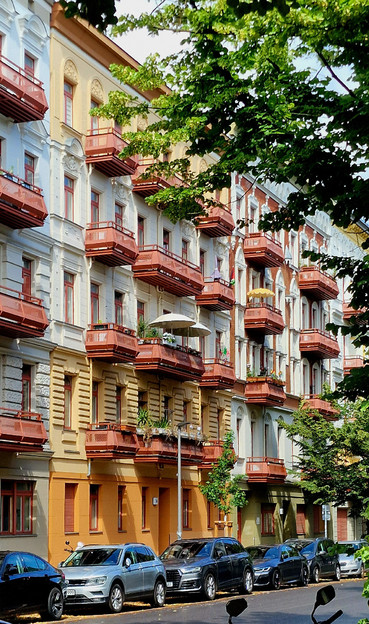 Mehrstöckiges Wohnhaus mit reich verzierten Fassaden und zahlreichen rotbraunen Balkonen in einer Baum gesäumten Straße im Berliner Stadtteil Wedding. Mehrere Autos parken am Straßenrand.


