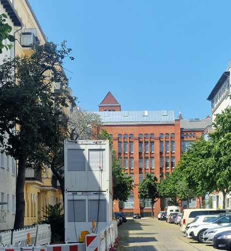 Blick auf eine gepflasterte Straße mit parkenden Autos und Baustellencontainern, im Hintergrund ein großes Backsteingebäude mit hohen Fenstern im Berliner Stadtteil Wedding.

