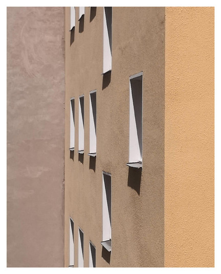 Fassade eines Wohnhauses mit beiger und gelber Putzstruktur; mehrere weiße, schräg geöffnete Fenster werfen lange Schatten.

