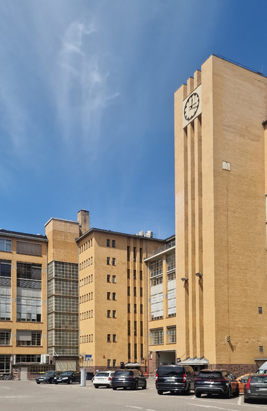 Backsteinbau im Stil der Moderne mit markantem Uhrenturm, großen Fensterfronten und geparkten Autos im Vordergrund auf einem Hofgelände im Berliner Bezirk Wedding.

