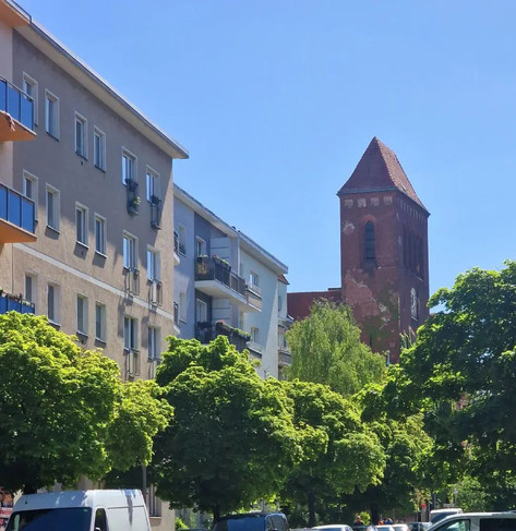 Straßenszene mit mehrgeschossigen Wohnhäusern, dichtem Baumbestand und dem Backsteinturm der Osterkirche im Berliner Stadtteil Wedding im Hintergrund.

