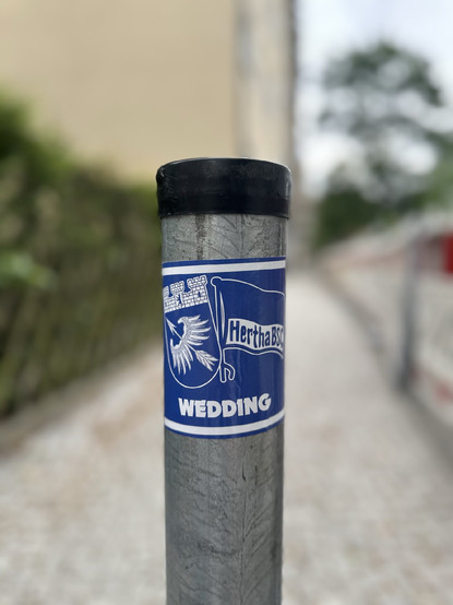 Nahaufnahme eines Aufklebers an einem Metallpfosten in Berlin-Wedding; der blaue Aufkleber zeigt das Logo von Hertha BSC, eine stilisierte Flagge, sowie ein Wappentier und den Schriftzug „WEDDING“.

