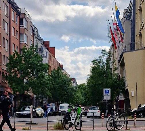 Straßenszene in Berlin-Wedding mit parkenden Autos, zwei Leihfahrrädern im Vordergrund und mehreren wehenden Fahnen an einem Hotelgebäude auf der rechten Seite. Die Straße ist von Bäumen gesäumt, bei bewölktem Himmel.

