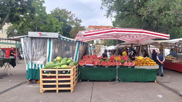 Marktstand auf dem Wochenmarkt mit gestreiftem Sonnendach, auf dem aufgeschnittene Wassermelonen, ganze Wassermelonen und gelbe Melonen verkauft werden; im Hintergrund weitere Stände und Passanten.

