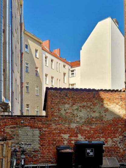 Hinterhof mit einer verwitterten roten Ziegelmauer, zwei Mülltonnen der BSR und abgestellten Fahrrädern; im Hintergrund mehrgeschossige Wohnhäuser mit Putzfassaden und rotem Ziegeldach unter klarem, blauem Himmel.

