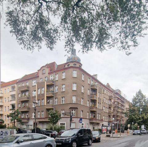 Straßenszene im Berliner Stadtteil Wedding mit einem fünfstöckigen Eckhaus im Gründerzeitstil, auffällig durch einen Turmaufsatz mit der Aufschrift „DW“; Balkone mit gelben Geländern, geparkte Autos und ein Baugerüst an der rechten Hausseite.


