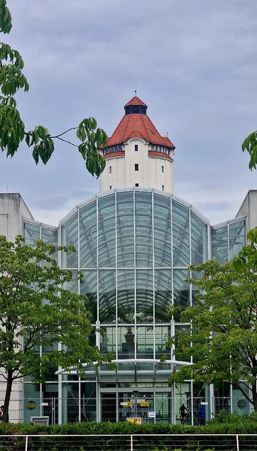 Glasüberdachter Eingang des Virchow-Klinikums in Berlin-Wedding mit dem historischen Wasserturm des Klinikums im Hintergrund, umgeben von Bäumen.


