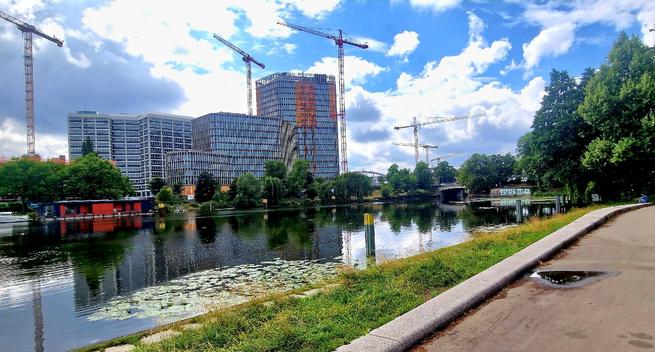 Blick auf den Berlin-Spandauer Schifffahrtskanal im Ortsteil Wedding mit modernen Bürogebäuden und mehreren Baukränen im Hintergrund; rechts eine Uferpromenade mit Bäumen.

