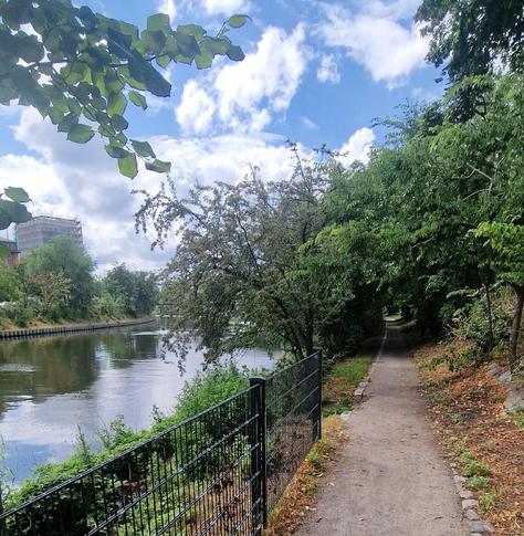 Uferweg am Berlin-Spandauer Schifffahrtskanal im Bezirk Wedding, rechts ein schmaler Fußweg unter Bäumen, links ein Zaun am Wasser mit Blick auf den Kanal und ein entferntes Gebäude mit Gerüst.

