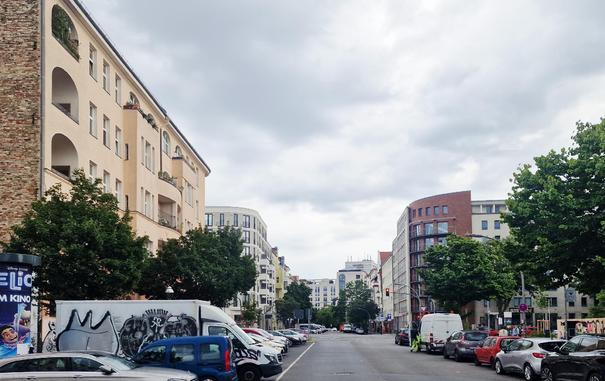 Straßenszene in Berlin-Wedding mit parkenden Autos, Wohnhäusern links und rechts der Fahrbahn und Bäumen am Straßenrand; im Hintergrund moderne und ältere Gebäude unter bewölktem Himmel.

