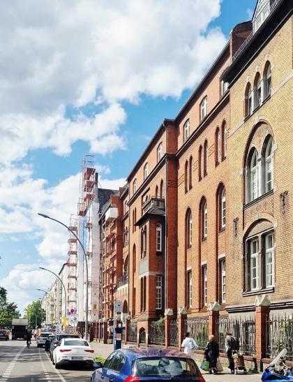 Backsteinfassade eines mehrstöckigen Altbaus in Berlin-Wedding bei Sonnenschein, mit Baugerüst an einem Nachbargebäude und parkenden Autos entlang der Straße.


