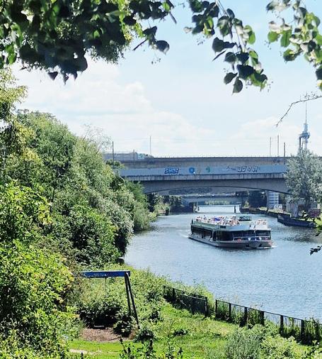Ausflugsboot auf dem Berlin-Spandauer Schifffahrtskanal in Wedding, umgeben von grüner Ufervegetation; im Hintergrund eine mit Graffiti bedeckte Brücke.


