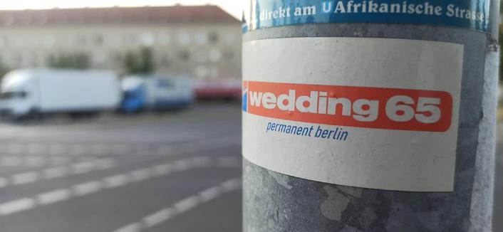 Aufkleber mit der Aufschrift „wedding 65 – permanent berlin“ an einem Laternenpfahl, im Hintergrund eine Straße mit parkenden Fahrzeugen im Berliner Stadtteil Wedding nahe der Afrikanischen Straße.

