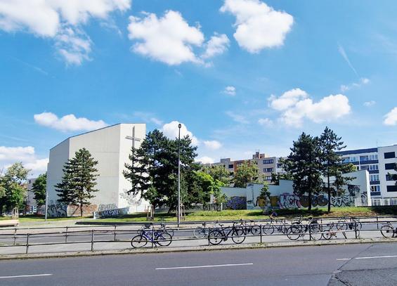 Weiße Kirche mit großem Kreuz an der Fassade in Berlin-Wedding, umgeben von Bäumen und Graffiti-besprühten Mauern; im Vordergrund eine Straße mit abgestellten Fahrrädern.

