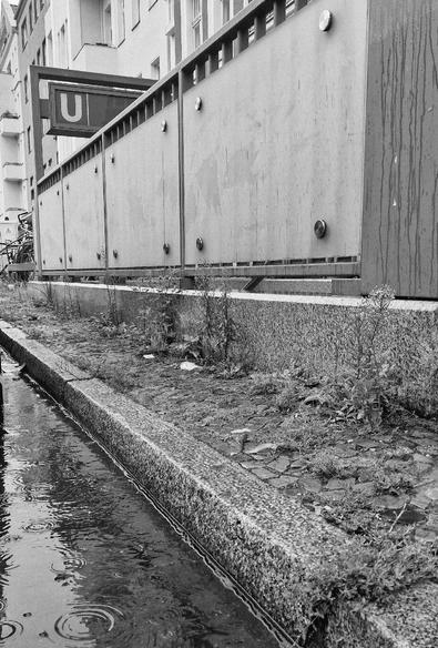 Schwarzweißfoto eines nassen Gehwegs mit Pfützen und Unkraut neben dem Eingang zur U-Bahn-Station Leopoldplatz in Berlin-Wedding.

