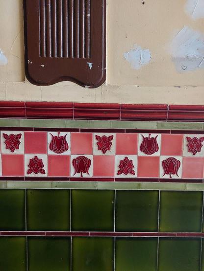 Fliesenwand mit floralen Jugendstil-Motiven: Obere Reihe aus rot-weißen Kacheln mit stilisierten Blütenmustern, darunter grüne, glänzende Kacheln in rechteckiger Anordnung.

