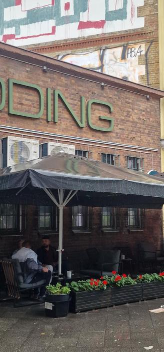 Zwei Männer sitzen unter einem großen schwarzen Sonnenschirm vor einem Backsteingebäude mit der Aufschrift „WEDDING“. Vor ihnen stehen Blumenkübel mit roten Geranien.

