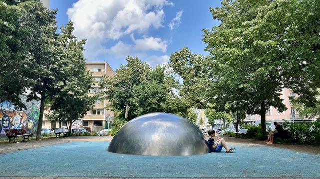 Halbkugelförmige, silberne Metallskulptur auf blauem Bodenbelag in einem kleinen Park in Berlin-Wedding, umgeben von Bäumen, Bänken und Wohnhäusern; ein Kind sitzt daneben und schaut auf ein Gerät.

