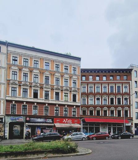 Altbaufassaden an der Müllerstraße mit Erdgeschossläden, darunter ein Reisebüro, ein Imbiss und ein Supermarkt; davor parkende Autos und eine Verkehrsinsel.


