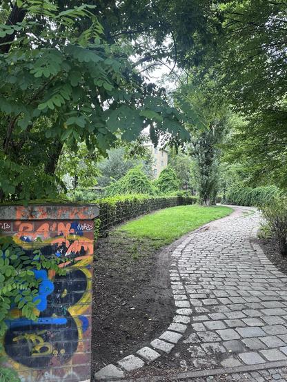 Gepflasterter Weg in einem parkähnlichen Bereich mit Bäumen und Sträuchern; links im Vordergrund eine mit bunter Graffiti besprühte Mauer, im Hintergrund ein mehrstöckiges Wohnhaus.

