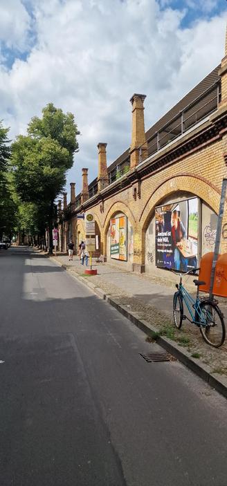 Straße mit Gehweg entlang eines gelben Backsteinviadukts mit Bögen und Plakaten; rechts ein abgestelltes Fahrrad, links Bäume und eine Bushaltestelle mit wartenden Personen.

