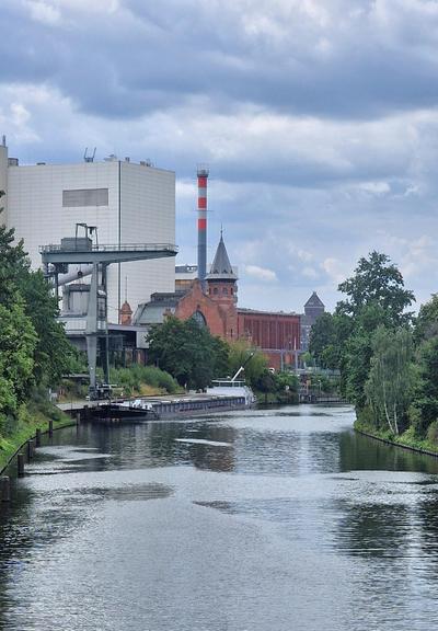 Blick auf den Berlin-Spandauer Schifffahrtskanal mit einem Frachtschiff am Ufer, links Industriegebäude mit Verladeanlage und rot-weißem Schornstein, im Hintergrund ein roter Backsteinbau mit Turm.

