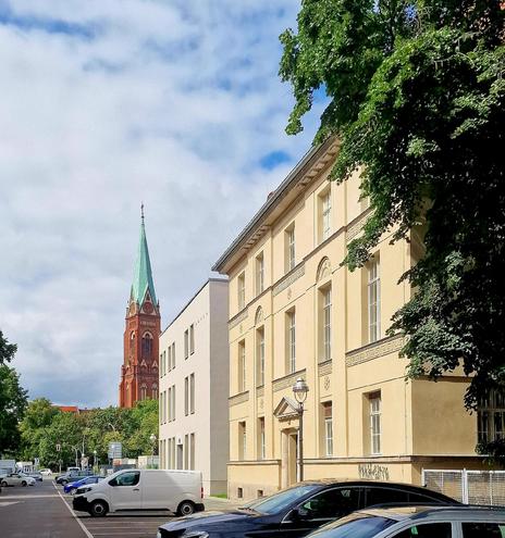 Straßenszene mit geparkten Autos, einem hellgelben Altbau rechts im Bild und der roten Backsteinkirche St. Joseph mit grünem Turmhelm im Hintergrund.

