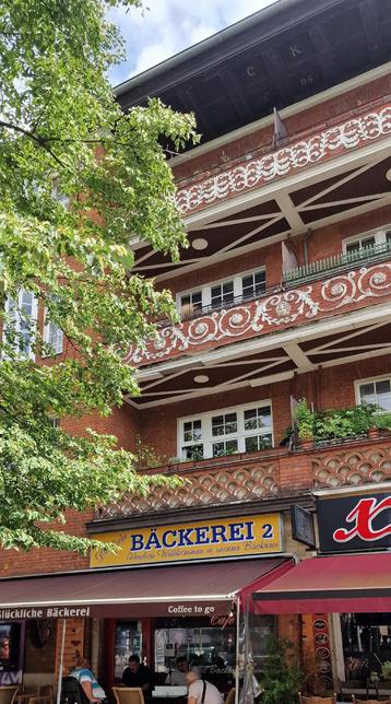Blick auf ein rot verklinkertes Wohnhaus mit verzierten Balkonen und dem Schriftzug „Bäckerei 2“ über einem Straßencafé im Erdgeschoss in Berlin-Wedding.

