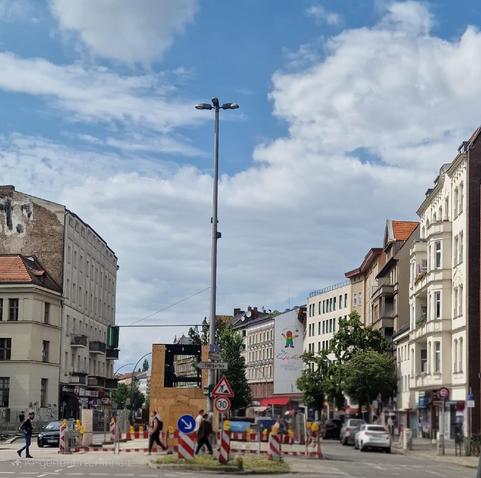 Straßenszene in Berlin-Wedding mit Baustellenabsperrungen, Fußgängern und Altbauten; im Hintergrund ein Wandbild mit der Aufschrift „Leben in Vielfalt“.

