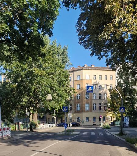 Straßenszene mit Fußgängerüberweg vor einem vierstöckigen Altbau in Berlin-Wedding, umgeben von Bäumen und mehreren Zebrastreifen-Schildern.

