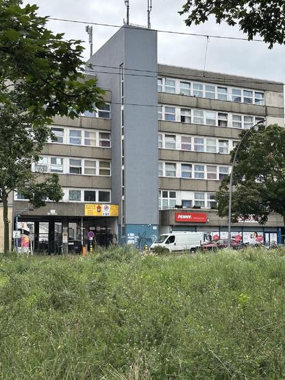 Wohnhaus mit grauer Fassade und zentralem Treppenhaus in Wedding, im Erdgeschoss ein PENNY-Supermarkt und ein Parkhauszugang; davor eine Wiese mit Wildwuchs.

