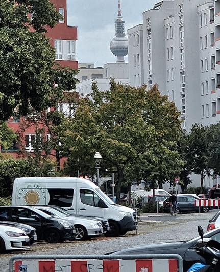 Blick auf einen Parkplatz mit mehreren Autos und einem Lieferwagen im Vordergrund, dahinter Bäume und Wohngebäude; im Hintergrund ist der Berliner Fernsehturm zwischen den Häusern sichtbar.

