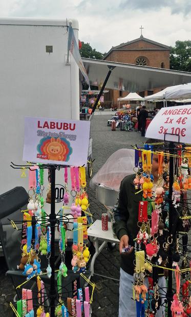 Ständer mit bunten Schlüsselanhängern auf einem Markt in Berlin-Wedding; im Hintergrund eine rote Backsteinkirche und Verkaufsstände.

