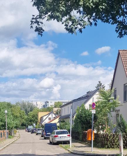 Das Bild zeigt eine ruhige Wohnstraße mit parkenden Autos am Straßenrand. Rechts stehen Einfamilienhäuser mit gepflegten Gärten, links eine Baum- und Heckenreihe entlang des Gehwegs. Ein Verkehrsschild weist auf ein eingeschränktes Halteverbot hin, daneben befindet sich ein orangefarbener Briefkasten. Im Hintergrund sind mehrstöckige Gebäude und üppiges Grün zu sehen, darüber ein teils bewölkter, teils blauer Himmel.

