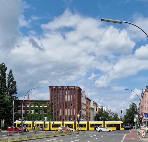 Gelbe Straßenbahn fährt an einer Kreuzung in Berlin-Wedding vor einem roten Backsteingebäude mit Spitzbogenfenstern entlang.

