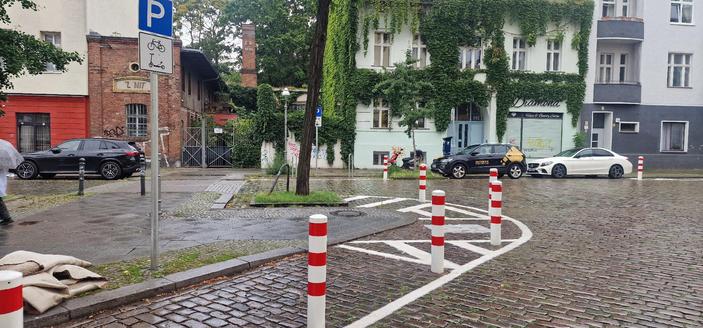 Nasse Kopfsteinpflasterstraße mit rot-weißen Pollern, parkenden Autos und einem grünen, mit Efeu bewachsenen Gebäude an einer Straßenecke in Berlin-Wedding.

