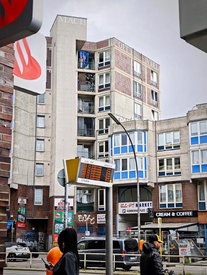 Wohnhaus am U-Bahnhof Pankstraße mit roter Kachelfassade und Schriftzug „Mach oft viel“ auf dem Dach, im Vordergrund Bushaltestelle mit digitaler Anzeigetafel und Passanten.

