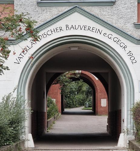 Bogenförmiger Durchgang mit der Aufschrift „Vaterländischer Bauverein e.G. gegr. 1902“, dahinter weitere Backsteinbögen und ein begrünter Weg.

