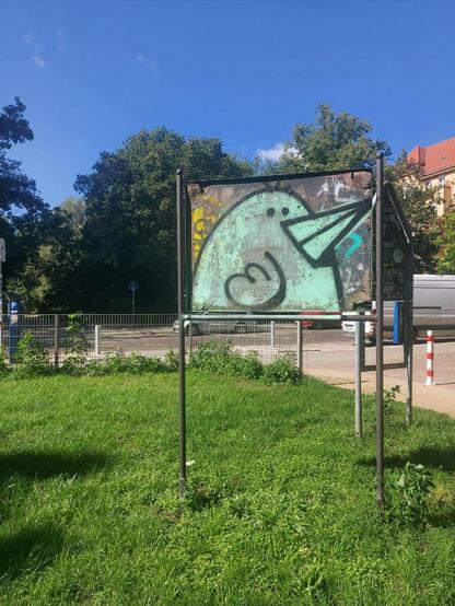 Verwittertes Schild mit einem Graffiti eines stilisierten Vogels in einem grünen Parkbereich in Berlin-Wedding.

