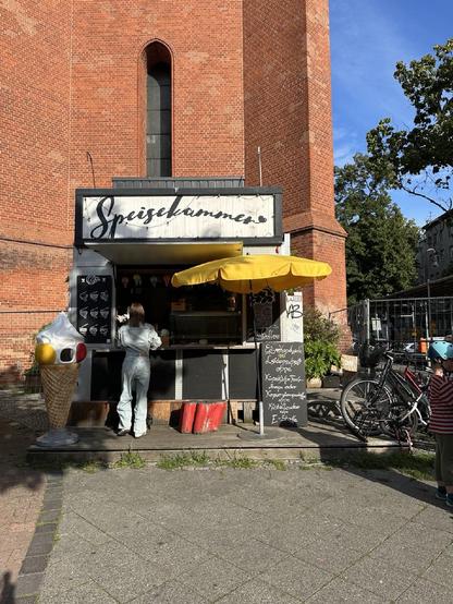 Das Foto zeigt einen kleinen Straßenverkaufsstand mit dem Namen „Speisekammer“, der sich direkt an einer roten Backsteinkirche befindet. Der Stand bietet offenbar Eis und Kaffee an, was durch die Eistüten-Dekoration und die Speisekarte mit Sorten wie „Eis Macadamia-Ladegeschmack“, „Käsekuchen-Birne“ oder „E-Stolle“ bestätigt wird. Eine Frau wird gerade bedient, während ein Kind mit Fahrradhelm in der Nähe steht. Die Szene wirkt freundlich und einladend bei sonnigem Wetter.

