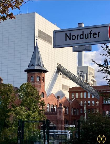 Straßenschild „Nordufer“ vor rotem Backsteingebäude mit Turm, im Hintergrund moderner, heller Industriebau mit Schornstein.

