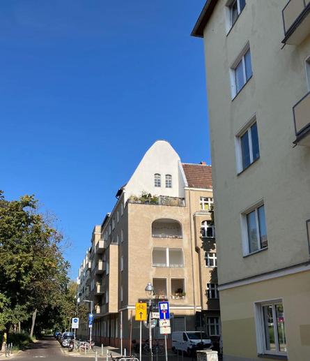Straßenszene mit mehrstöckigen Wohnhäusern, darunter ein Gebäude mit abgerundetem Giebel und Balkonen, bei klarem blauem Himmel.

