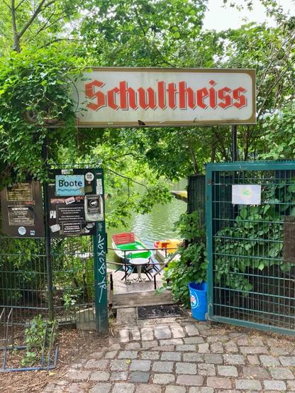 Tor mit Schild „Schultheiss“ führt zu einem kleinen Bootsanleger am Wasser, umgeben von dichtem Grün.

