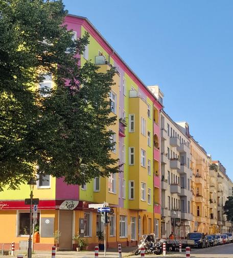 Mehrstöckiges Wohnhaus in Wedding mit farbiger Fassade in Gelb, Pink und Grün an einer Straßenecke, daneben weitere Häuser in helleren Farbtönen.

