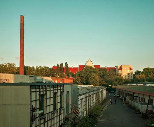Industriegelände mit Lagerhallen und hohem Schornstein, im Hintergrund Bäume und Gebäude mit roten Dächern.

