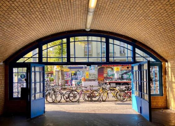 Blick durch ein gemauertes Tor mit blauen Türen auf eine Straße mit abgestellten Fahrrädern und einem bunt bemalten Lieferwagen.

