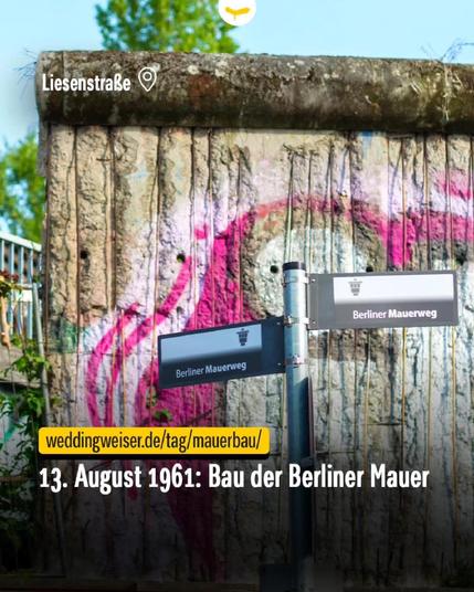 Fragment der Berliner Mauer mit pinkem Graffiti an der Liesenstraße, davor zwei Straßenschilder mit der Aufschrift „Berliner Mauerweg“.

