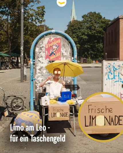 Straßenstand auf dem Leopoldplatz mit gelbem Sonnenschirm, an dem eine Person frische Limonade verkauft; handgemaltes Schild „Frische Limonade“ an der Tischfront.

