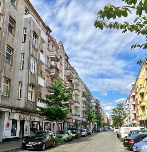 Blick auf eine Wohnstraße in Berlin-Wedding mit mehrstöckigen Altbauten, parkenden Autos am Straßenrand und einzelnen Bäumen, unter teils bewölktem Himmel.

