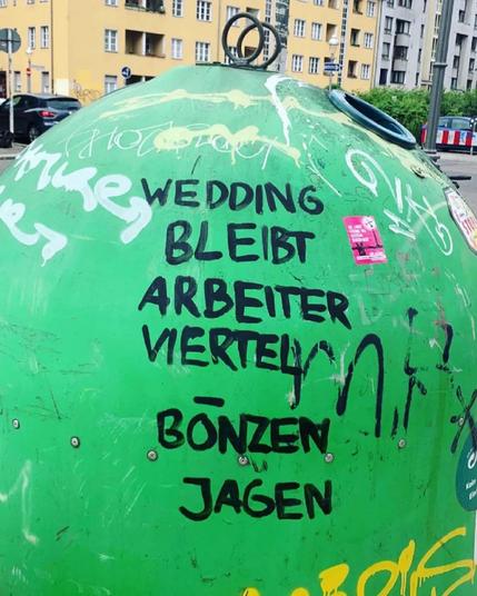 Grüner Altglascontainer mit schwarzem Graffiti-Schriftzug „Wedding bleibt Arbeiterviertel, Bonzen jagen“, umgeben von weiteren Kritzeleien, im Hintergrund Wohnhäuser.

